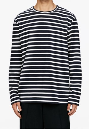 Persona che indossa una maglietta a righe orizzontali bianche e nere a maniche lunghe e pantaloni neri, in piedi davanti a uno sfondo bianco uniforme.