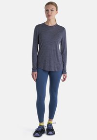 Langärmliges graues Sportoberteil, kombiniert mit blauen Leggings und schwarzen Laufschuhen. Glatte Textur, figurbetontes Design und abgerundeter Saum.