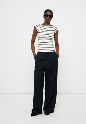 Femme portant des lunettes de soleil noires, un haut ajusté à manches courtes rayé blanc et noir, un pantalon noir à jambes larges et des chaussures noires à bouts pointus, debout les mains dans les poches.