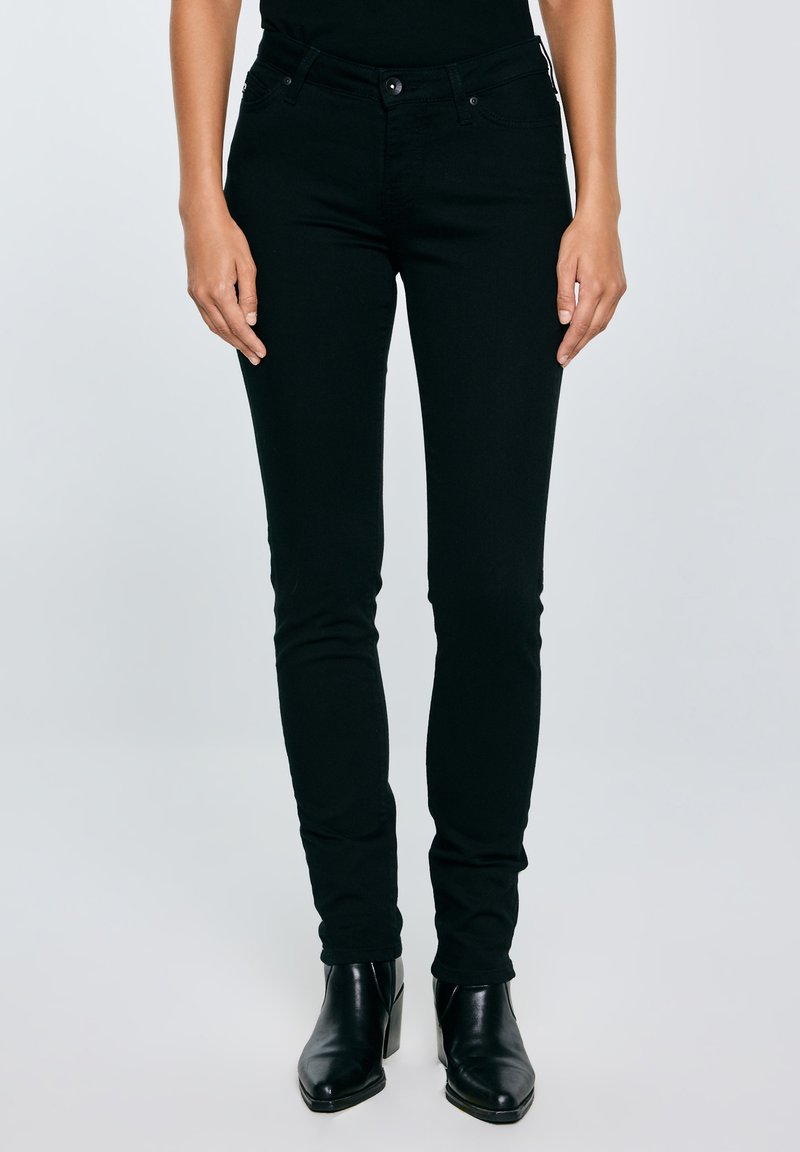 Nő fekete, testhezálló farmerben, fekete hegyes orrú bokacsizmában és fekete felsőben, egyszínű világosszürke háttér előtt állva.