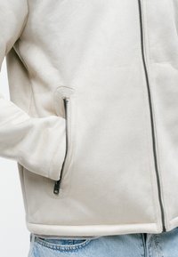 Giacca in suede beige con una texture liscia, caratterizzata da una zip frontale e tasche laterali con zip. Progettata con una silhouette aderente.