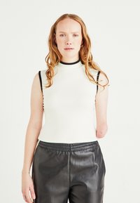 Young woman with wavy red hair wearing sleeveless white top and black pants, with left forearm amputated below elbow, standing against white background.