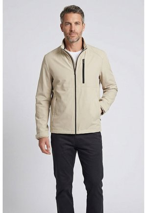 Man draagt beige jas met rits over wit shirt en zwarte broek, staat met één hand in de jaszak tegen een effen achtergrond.