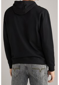 Sudadera negra con capucha de textura suave. Presenta un bolsillo tipo canguro y puños acanalados, combinada con unos jeans ajustados de color gris claro con costuras decorativas.
