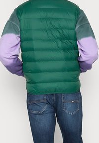 Man med en grön vadderad väst över en lila och grå långärmad skjorta, tillsammans med mörkblå jeans, vänd bort.