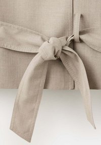 Ceinture en tissu beige nouée en un nœud avec des surpiqûres visibles sur un fond de vêtement beige texturé assorti.