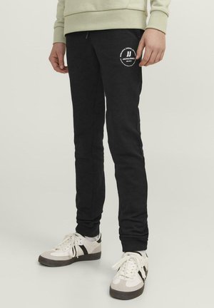 Pantalones de chándal negros de tejido suave, con cintura ajustable mediante cordón y un logo blanco en el muslo izquierdo. Estilo ajustado, con tobillos acampanados.