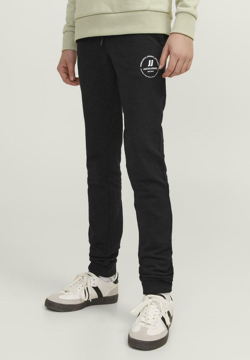 Pantalones de chándal negros de tejido suave, con cintura ajustable mediante cordón y un logo blanco en el muslo izquierdo. Estilo ajustado, con tobillos acampanados.
