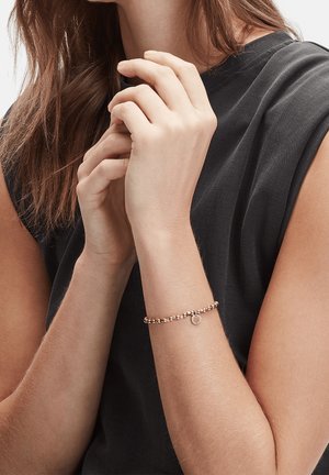 Femme portant un haut noir sans manches, aux longs cheveux bruns, joignant les mains près de la poitrine, portant un délicat bracelet en or avec des perles et un petit pendentif.