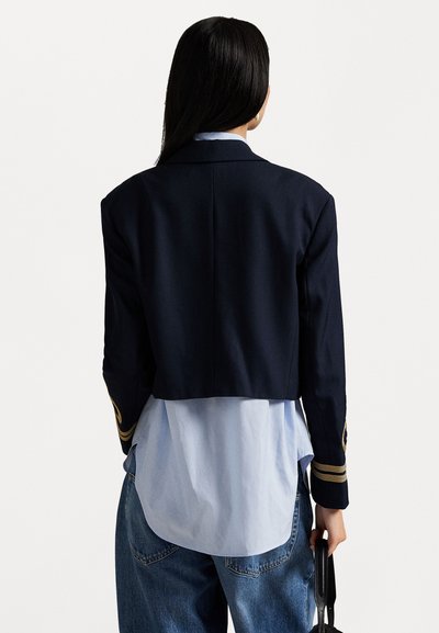 Cropped navy blazer with gold accents and structured shoulders, worn over a light blue shirt and paired with loose denim jeans.