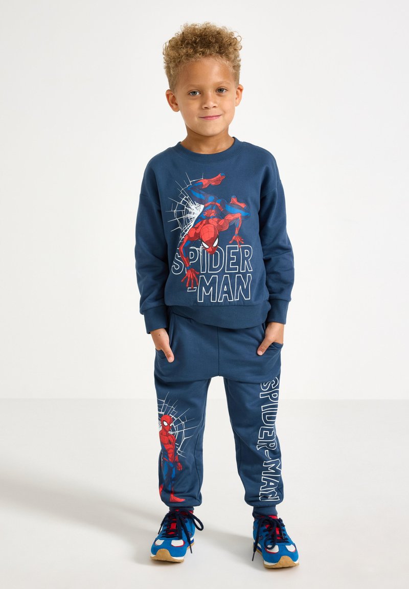 Nuori poika, jolla on kiharat hiukset, pukeutuneena siniseen Spider-Man-huppariin ja samansävyisiin housuihin, seisoo kädet taskuissa valkoista taustaa vasten.