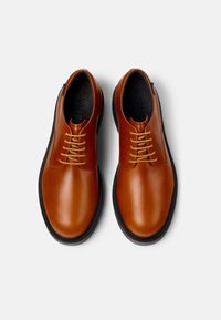 Zapatos de cuero marrón con una textura suave, con punteras redondas, un perfil bajo y cordones de color marrón claro. Se pueden ver las suelas de goma negras.