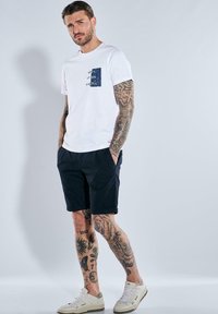 White cotton t-shirt with a blue patterned pocket, paired with navy shorts and cream sneakers, featuring various tattoos on arms and legs.