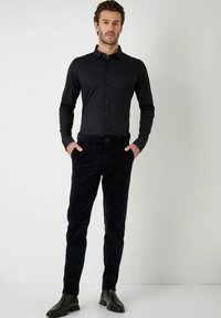 Marineblauw overhemd met knopen en een subtiel stippenpatroon, gecombineerd met donkere corduroy broek en zwarte chelsea boots, staande tegen een effen achtergrond.