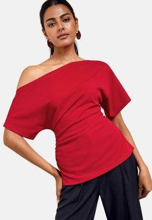 Kvinde iført en rød off-shoulder top med korte ærmer og sorte bukser, poserer med den ene hånd på hoften mod en ensfarvet baggrund.