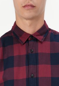 Karohemd in Rot- und Marineblau mit Kragen, aus glattem Stoff und schwarzen Knöpfen. Nahaufnahme des Brustbereichs.