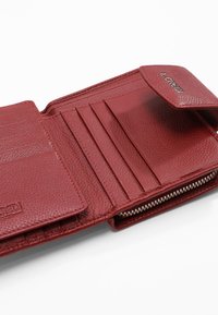 Portefeuille en cuir rouge avec une finition texturée, comportant des emplacements pour cartes et une poche zippée. Rabat de fermeture avec un détail de logo en relief.
