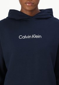 Námornícky modrá mikina vyrobená z bavlny, s predným kengurím vreckom a bielym nápisom „Calvin Klein“ potlačeným na hrudi.