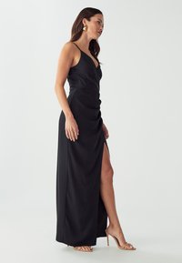 Schwarzes Satin-Maxikleid mit tiefem V-Ausschnitt, geraffter Front und seitlichem Schlitz bis zum Oberschenkel. Model trägt beige High-Heels und auffällige Ohrringe.