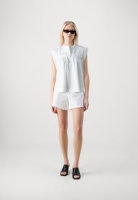 Gaiši zils bez piedurknēm topiņš ar teksturētu audumu un priekšējiem pogām, apvienots ar baltām izsistām šortiem un melniem atvērtajiem zābakiem.