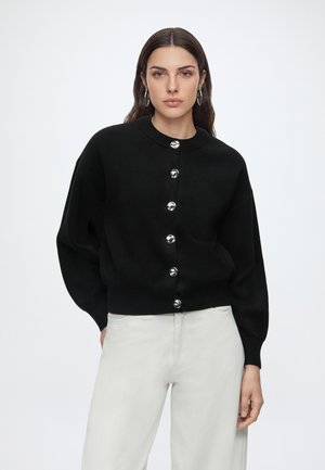 Femme aux longs cheveux foncés porte un cardigan noir boutonné et un pantalon blanc, debout devant un fond clair uni.