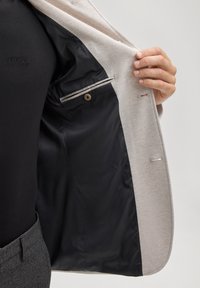 Blazer grigio chiaro con un tessuto esterno liscio, fodera in raso nero, chiusura con un solo bottone e tasca interna con dettaglio a bottone.