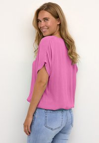 Løs pasform, kortærmet top i levende pink stof. Har en let tekstur og en buet kant, parret med lyseblå jeans.