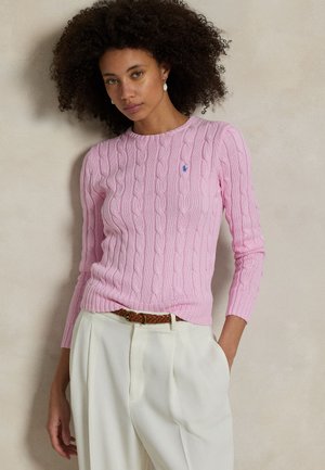 Femme aux cheveux bouclés portant un pull en maille torsadée rose et un pantalon blanc avec une ceinture tressée marron sur un fond neutre.