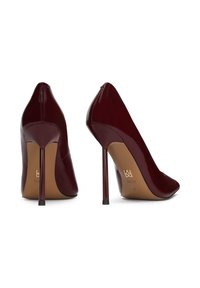 Tmavě červené lakované lodičky s vysokými podpatky a špičatou špičkou, s jehlovými podpatky a béžovými podrážkami, na kterých je viditelné logo značky.