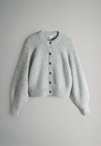 Gaiši pelēks adīts kardigans ar ribotu tekstūru, apaļu apkakli un piecām bronzas pogām priekšpusē. Tam ir balonu formas piedurknes un īsāks garums.