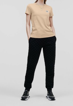 Kobieta ubrana w beżowy t-shirt z krótkim rękawem, czarne joggery we wzory i czarne trampki z białą podeszwą, stoi z rękami w kieszeniach.
