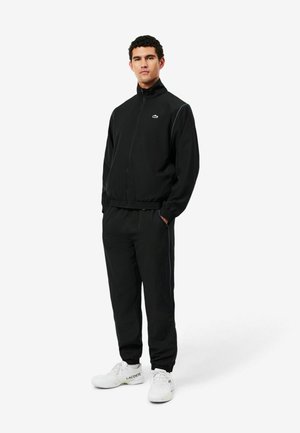 Uomo in piedi con le mani in tasca che indossa una giacca da jogging nera con zip e pantaloni con sneakers bianche Lacoste su sfondo bianco.