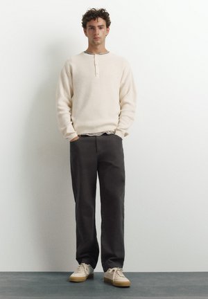 Ung mand stående med hænderne i lommerne iført en cremefarvet langærmet henley, mørke bukser og beige sneakers mod en ensfarvet baggrund.