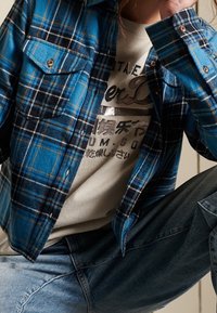 Camicia di flanella a quadri blu e nera con due tasche sul petto e chiusura a bottoni, indossata sopra una maglietta grigia con grafica. Jeans in denim visibili.