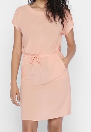 Frau trägt ein hellrosa kurzärmeliges Kleid mit Taillenbindeband, eine Hand in der Tasche, steht vor weißem Hintergrund.