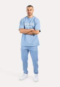 Hellblaues Sportset, bestehend aus einem T-Shirt und einer Sweatpants, mit einem Logo auf der Brust und dem linken Oberschenkel, kombiniert mit weißen Sneakers.