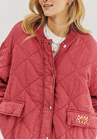 Prešívaná červená bunda s patentkami, s diamantovým vzorom, dvoma prednými vreckami a malou kockovanou nášivkou s grafickým dizajnom.