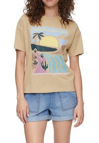 Beige Baumwoll-T-Shirt mit einem Grafikdruck, der einen Sonnenuntergang, eine Palme und eine Strandlandschaft in Pastellfarben zeigt. Kombiniert mit Denim-Shorts.