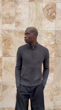 Ein Mann mit kurzem blonden Haar, der einen dunkelgrauen Reißverschluss-Pullover und schwarze Hosen trägt, steht an einer beige marmorierten Wand und schaut nach links.