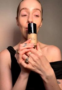 Person with slicked-back hair and rings holds a bottle of luminous silk foundation close to their face against a plain background.