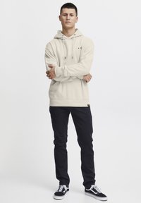 Beige hooded sweatshirt with a front pocket and drawstring, paired with dark slim-fit jeans and black sneakers. Simple, casual design.