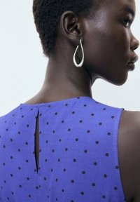 Purple fabric top with black polka dots, featuring a back keyhole detail. Silver teardrop earrings complement the design.