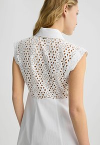 Camicia bianca con colletto, dotata di un retro in broderie anglaise. Le cuciture a vista e le maniche corte mettono in risalto il motivo simile al pizzo.