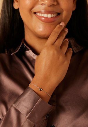 Silbernes Armband mit einem herzförmigen Anhänger, getragen an einer Hand, die auf einem braunen Satinkleid ruht. Glatte Textur, schlichtes Design.