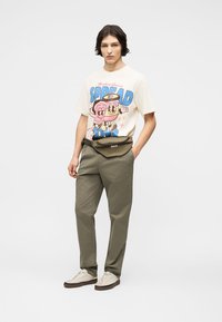 Νέος άνδρας που φοράει μπλουζάκι με το γραφικό "Spread Joys", ελαιδί παντελόνι, μπεζ αθλητικά παπούτσια και μία ελαιδί τσάντα μέσης, στέκεται με τα χέρια στις τσέπες.