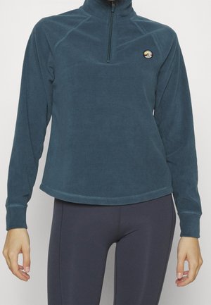 Person trägt einen türkisen Fleecetop mit Reißverschluss bis zur Brust und dunkle Leggings, steht vor einem schlichten hellen Hintergrund.