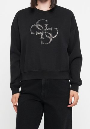 Person iført en sort sweatshirt med et stort sølvfarvet mønstret overlappende bogstavlogo på brystet, kombineret med sorte bukser.