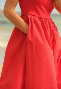 Robe rouge sans manches avec un corsage ajusté et une jupe évasée dotée de poches, fabriquée dans un tissu lisse qui tombe gracieusement.