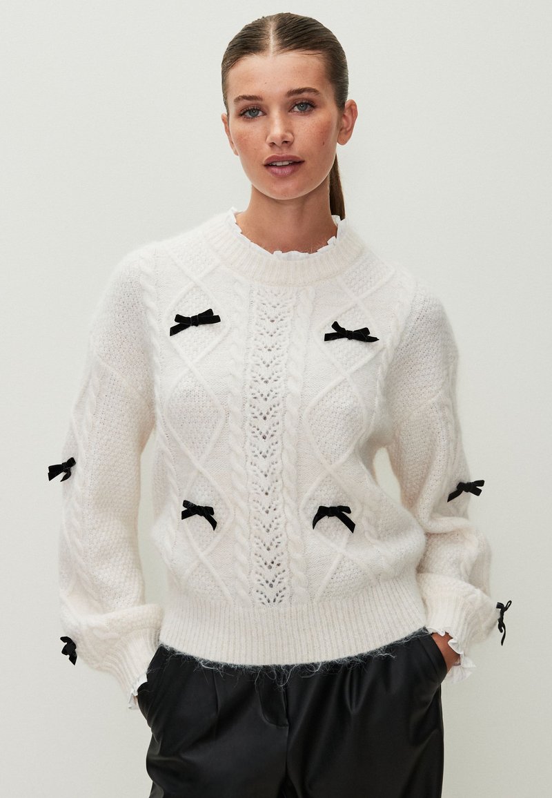 Suéter blanco de punto con un patrón de diamantes, textura en relieve, acentos de cinta negra en las mangas y un cuello acanalado.