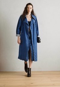 Trench coat in denim con design doppiopetto, caratterizzato da maniche arrotolate e uno spacco frontale, abbinato a stivaletti neri alla caviglia e una borsa piccola.